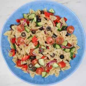 A colorful Mediterranean pasta salad with cherry tomatoes, cucumbers, olives, and bell peppers in a blue bowl.