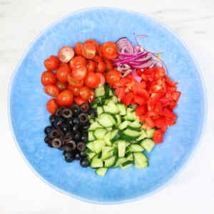 A colorful arrangement of chopped vegetables including tomatoes, cucumbers, sweet peppers, red onions, and black olives.