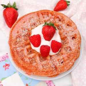 A pink waffle topped with whipped cream and fresh strawberries, dusted with powdered sugar.