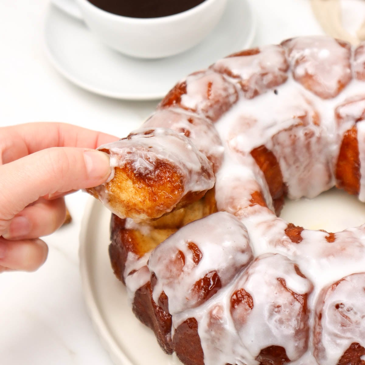 A hand pulls apart a piece of glazed monkey bread from a circular loaf.