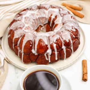 A glazed pull-apart monkey bread on a plate with a cup of coffee and cinnamon sticks nearby.
