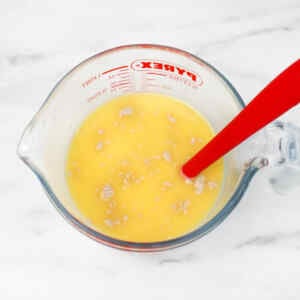 A measuring cup filled with a yellow mixture of milk, water, sugar, melted butter, yeast, and a red spoon for stirring it.