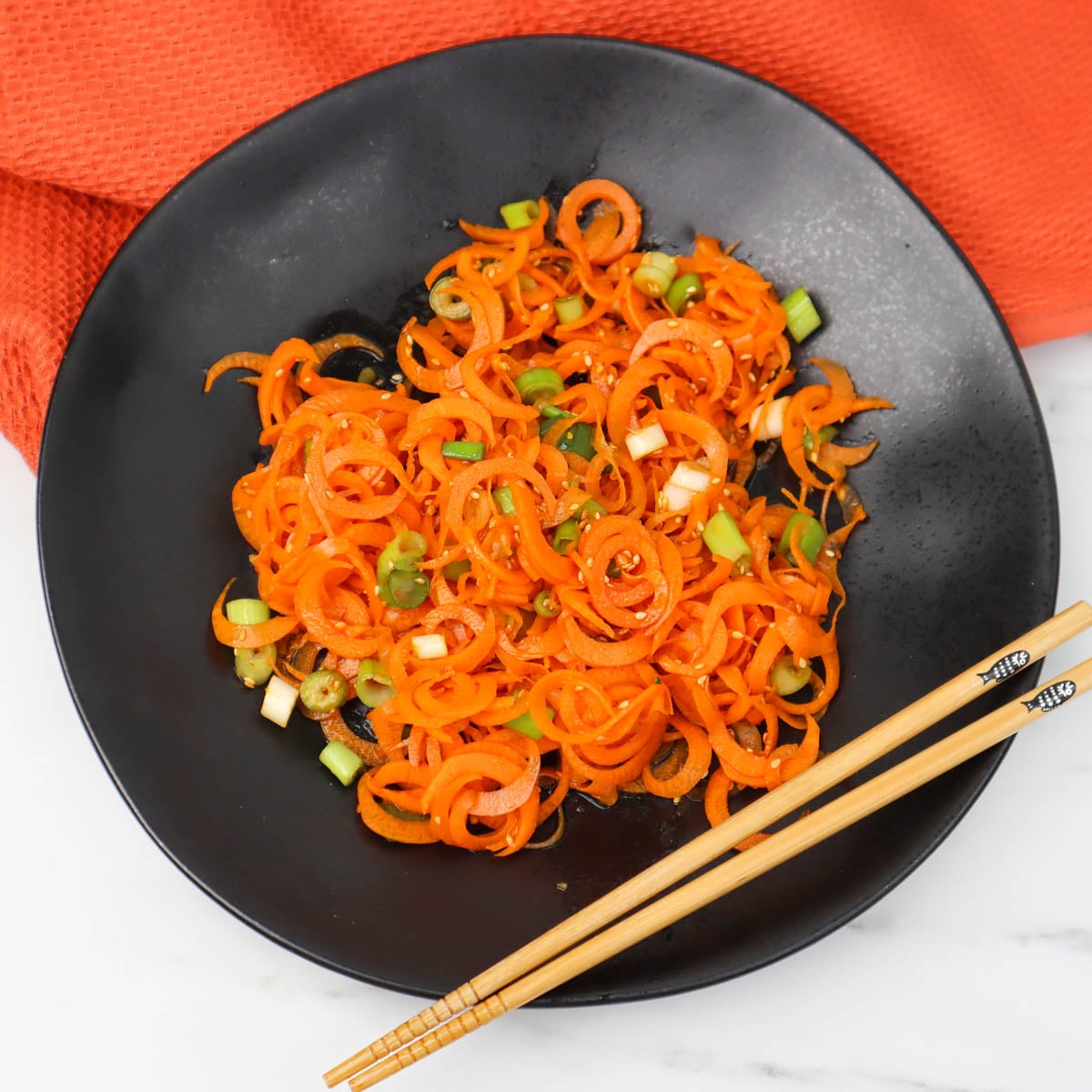 Asian carrot salad A black plate filled with spiralized carrot salad topped with green onions and served with chopsticks.