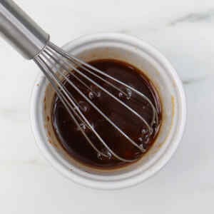 A whisk mixing a dark sauce in a small white bowl.