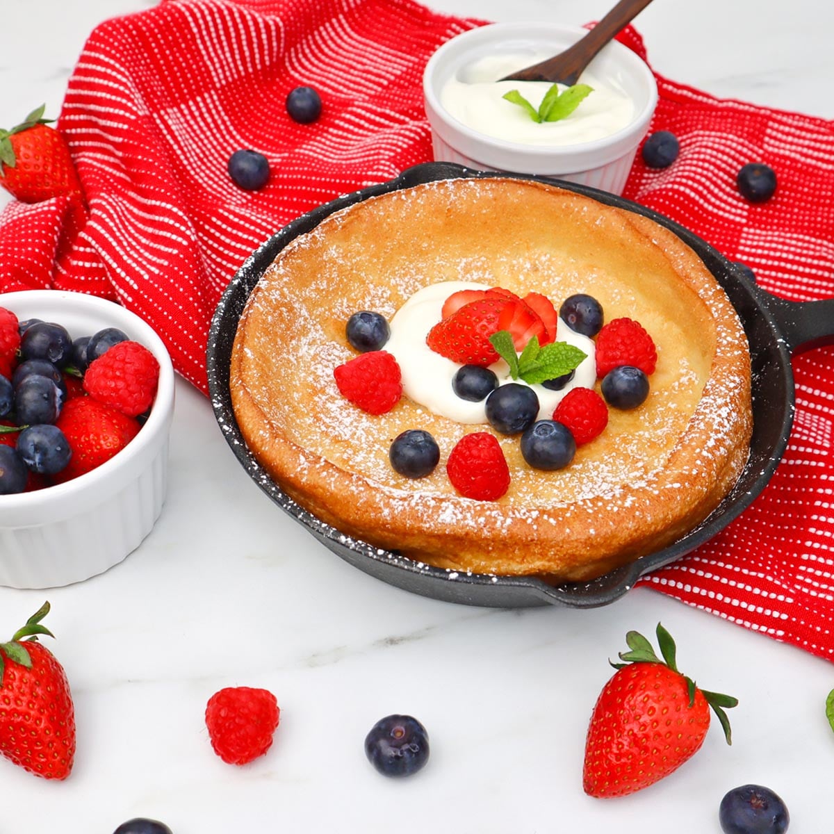 Dutch Baby Pancake Ein Dutch Baby Pancake mit Beeren und Sahne, serviert in einer gusseisernen Pfanne auf einem rot-weiß gestreiften Handtuch.