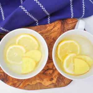 Duas xícaras de chá de limão e gengibre com fatias de limão e gengibre sobre uma superfície de madeira.