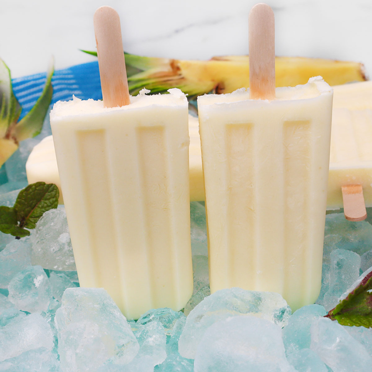 Pineapple Popsicles Two pineapple popsicles on ice with mint leaves and pineapple pieces in the background.