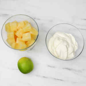 Chopped pineapple in a bowl, a lime, and a bowl of creamy mixture on a marble surface.