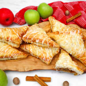 Golden puff pastry apple turnovers drizzled with icing, surrounded by apples and cinnamon sticks.