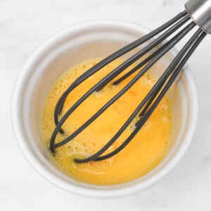 A whisk mixing beaten eggs in a small white bowl.