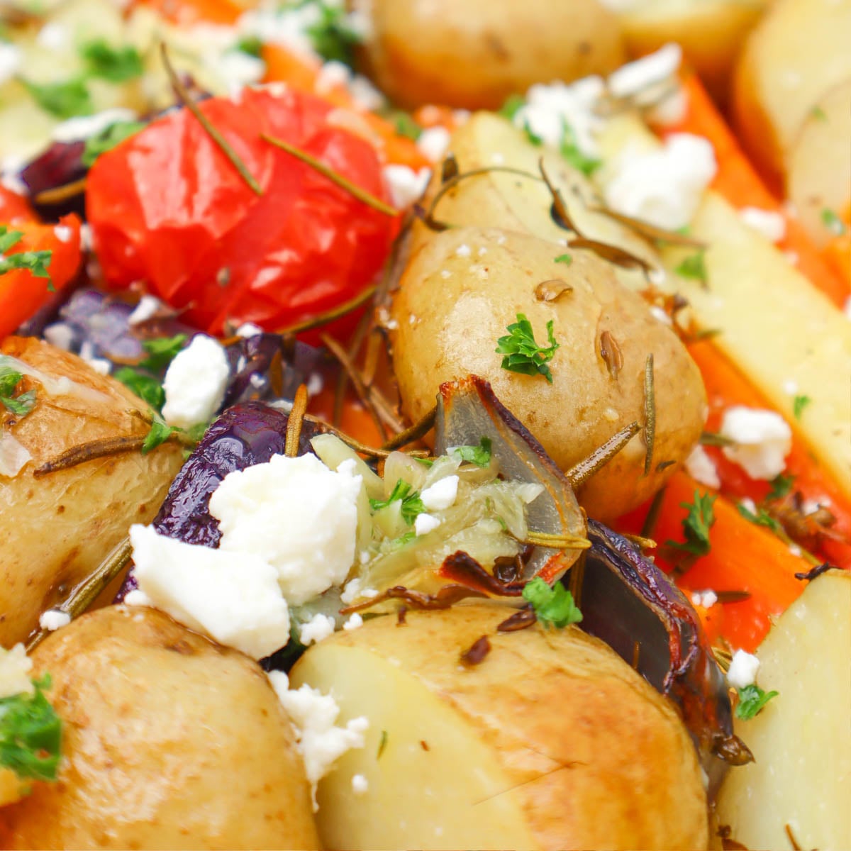 Roasted Vegetable Tray Bake A close-up of roasted vegetables including potatoes, carrots, and tomatoes topped with feta cheese and herbs.