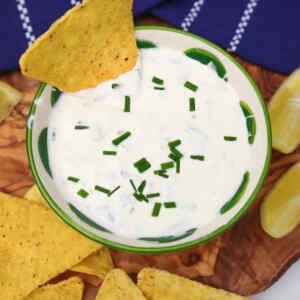 A bowl of sour cream and chive dip garnished with chives, surrounded by tortilla chips and lemon wedges.