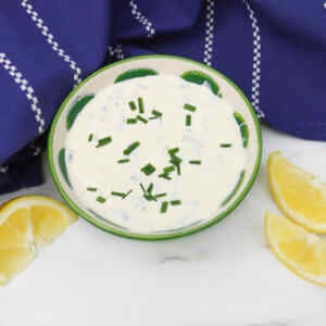 A bowl of sour cream and chive dip garnished with chopped chives, next to lemon wedges.