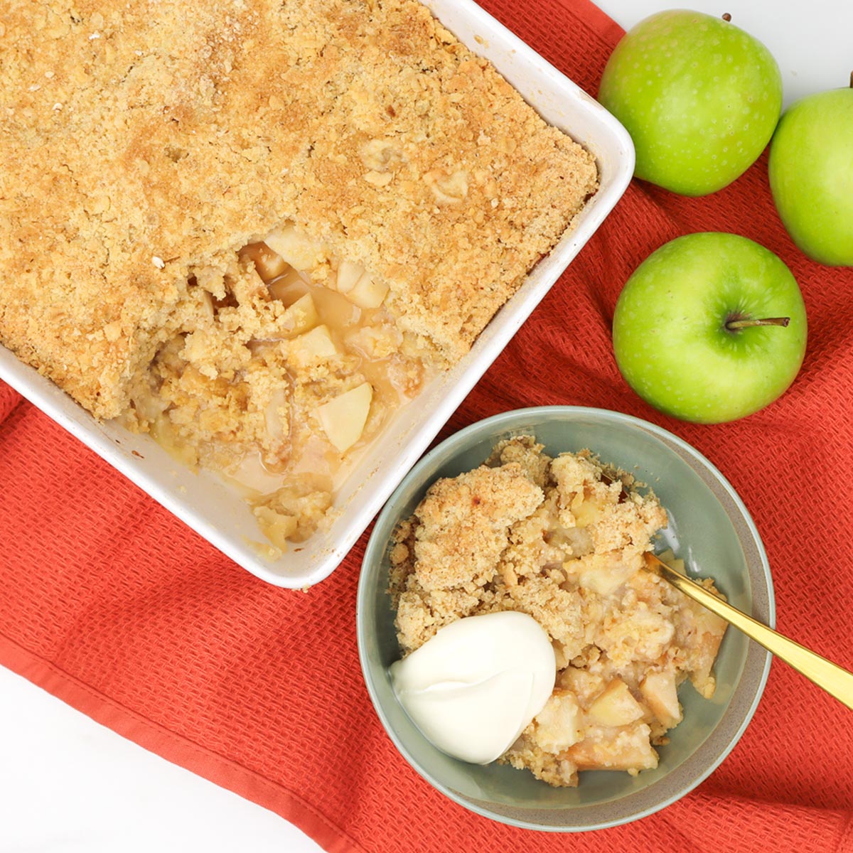 Apple Crumble A serving of apple crumble with a scoop of cream beside a baking dish and green apples.