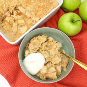Apple crumble in a bowl topped with cream and a serving dish with apple crumble ready to be served