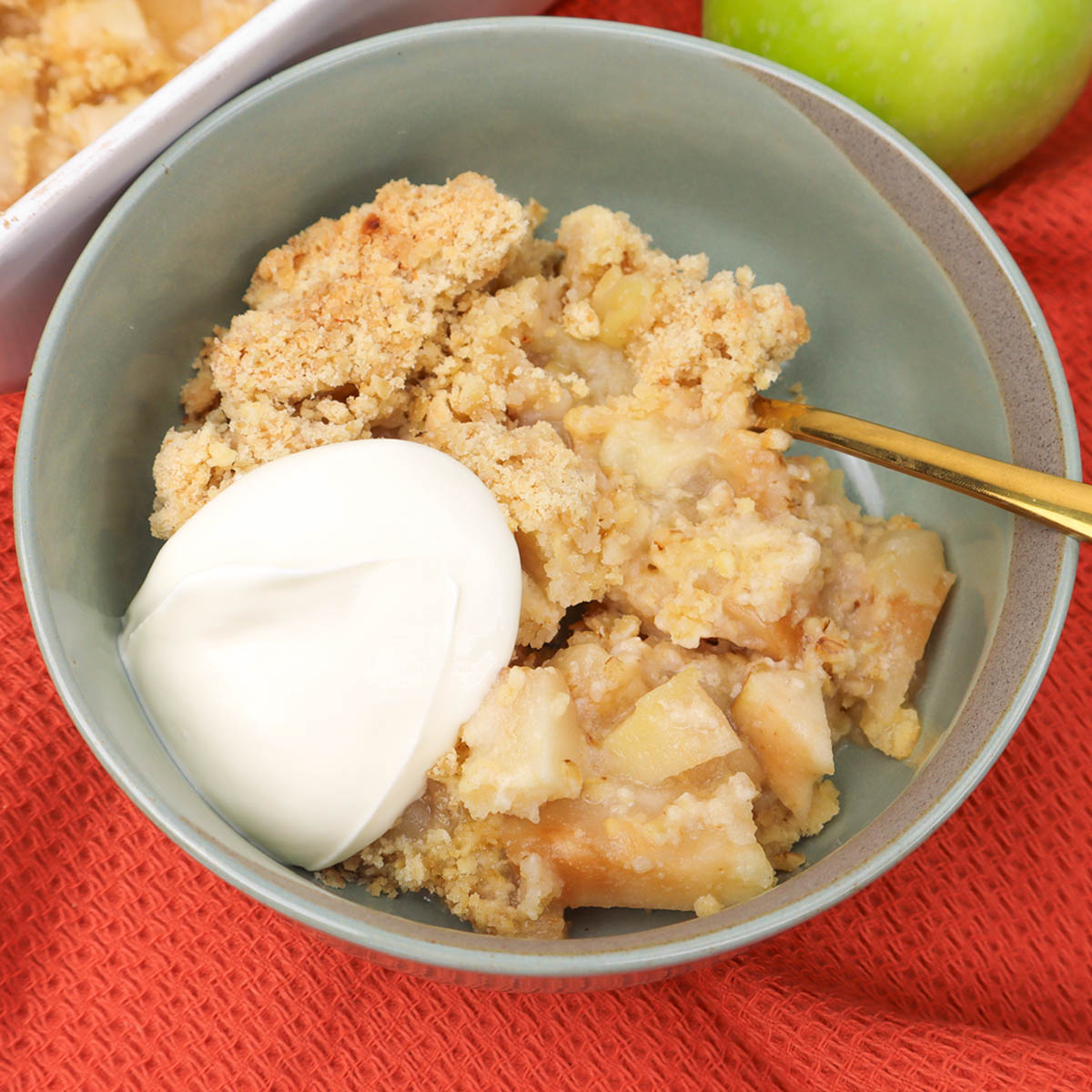 Apple Crumble A bowl of apple crumble topped with a dollop of cream on a textured red surface.
