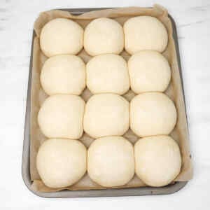 Twelve uncooked burger buns arranged on a baking tray lined with parchment paper.