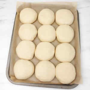 Twelve round, uncooked burger buns arranged on a baking tray lined with parchment paper.
