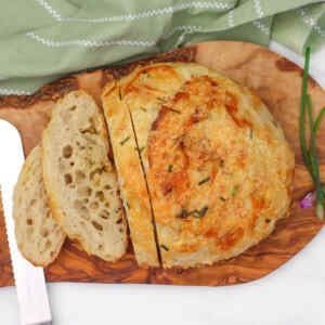 A sliced cheese and chive sourdough loaf on a wooden board with a knife and chives nearby.