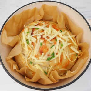 A rustic cheese and chive sourdough loaf topped with shredded cheese and chives, resting in a baking pot.