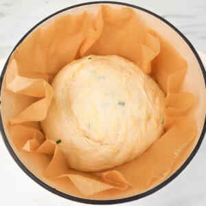 A round dough with cheese and chives resting in a pot lined with parchment paper.
