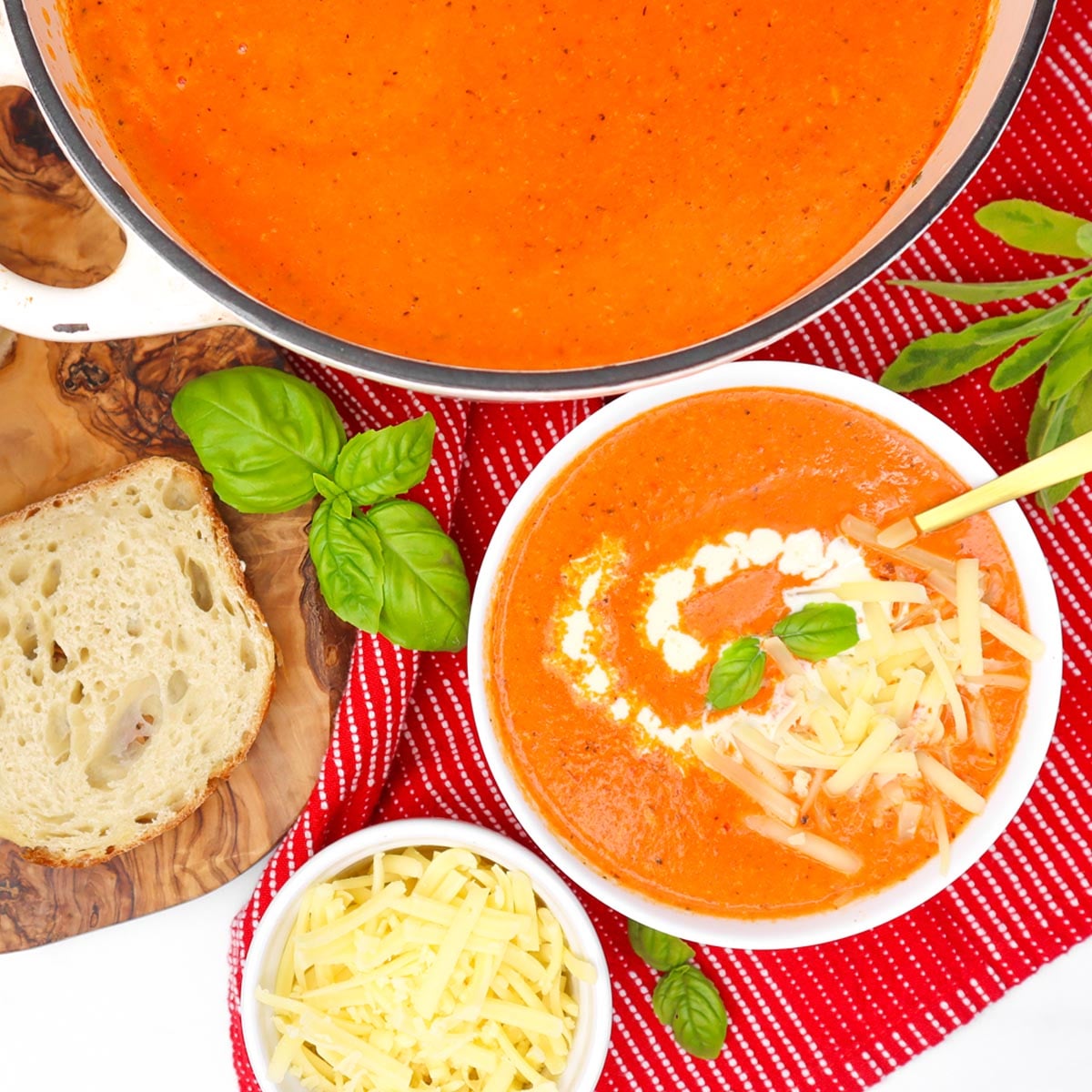 Cheesy Roasted Red Pepper Soup A bowl of cheesy roasted red pepper soup garnished with cream and basil, alongside a slice of bread and shredded cheese.
