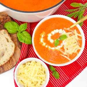A bowl of cheesy roasted red pepper soup garnished with basil and cheese, alongside a slice of bread.