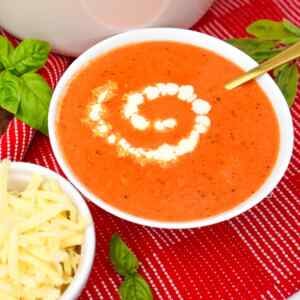 A bowl of cheesy roasted red pepper soup garnished with a swirl of cream, alongside a bowl of shredded cheese and fresh basil.