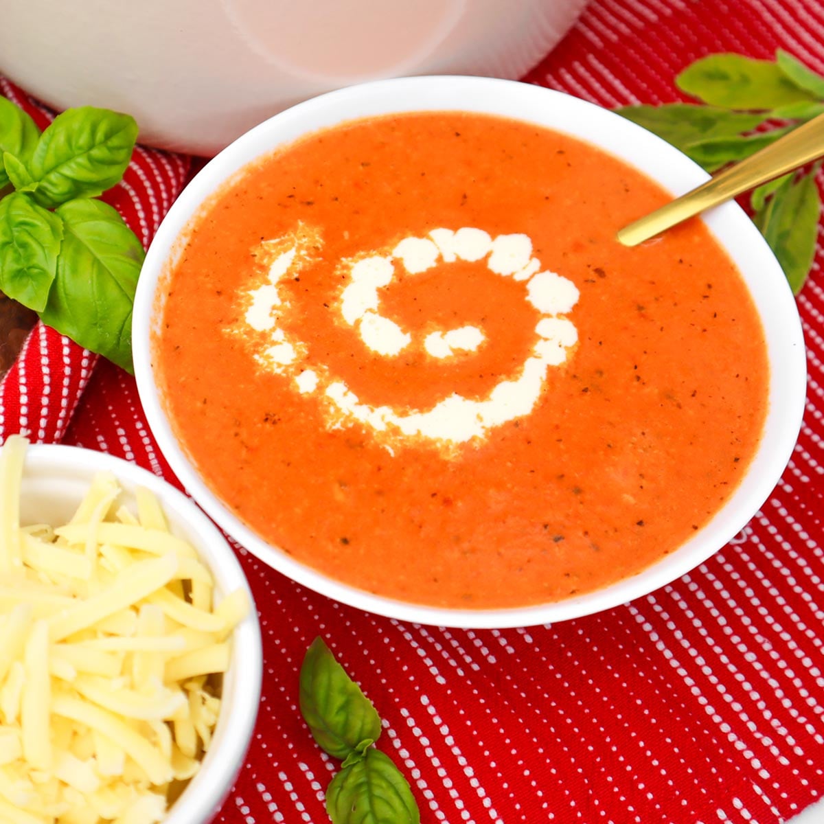 Cheesy Roasted Red Pepper Soup A bowl of cheesy roasted red pepper soup garnished with a swirl of cream, alongside a bowl of shredded cheese and fresh basil.