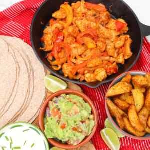 A skillet with chicken fajitas alongside tortillas, guacamole, and seasoned potato wedges.