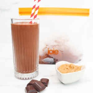 A glass of chocolate peanut butter smoothie with a striped straw, chocolate pieces, and peanut butter in a bowl.
