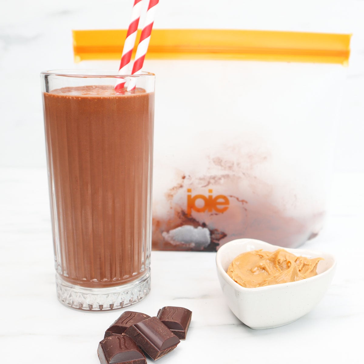 Chocolate Peanut Butter Smoothie Freezer Meal Prep Packs A glass of chocolate peanut butter smoothie with a striped straw, chocolate pieces, and peanut butter in a bowl.