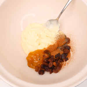 A bowl containing mayonnaise, spices, raisins, and a spoon mixing the ingredients.