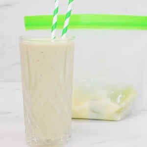 A glass of kiwi and avocado smoothie with green striped straws next to a freezer bag.