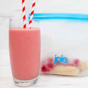 A glass of raspberry banana smoothie with red and white striped straws next to a freezer bag of ingredients.