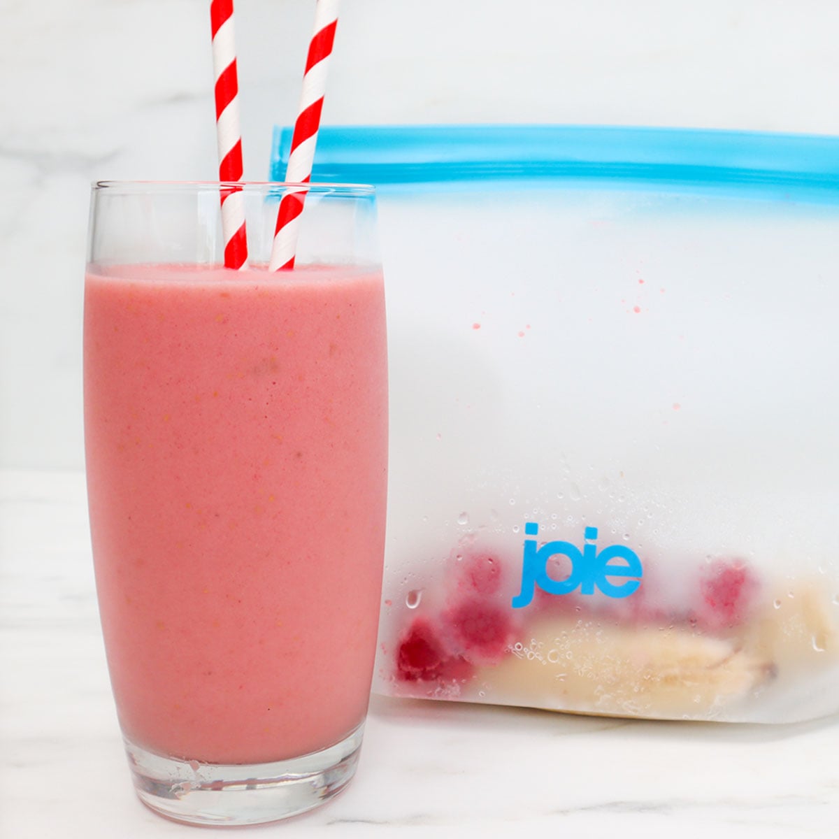 Raspberry Banana Smoothie Freezer Meal Prep Packs A glass of raspberry banana smoothie with red and white striped straws next to a freezer bag of ingredients.