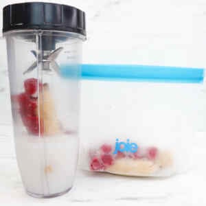 A blender filled with raspberries, banana, and milk next to a freezer bag with smoothie ingredients.