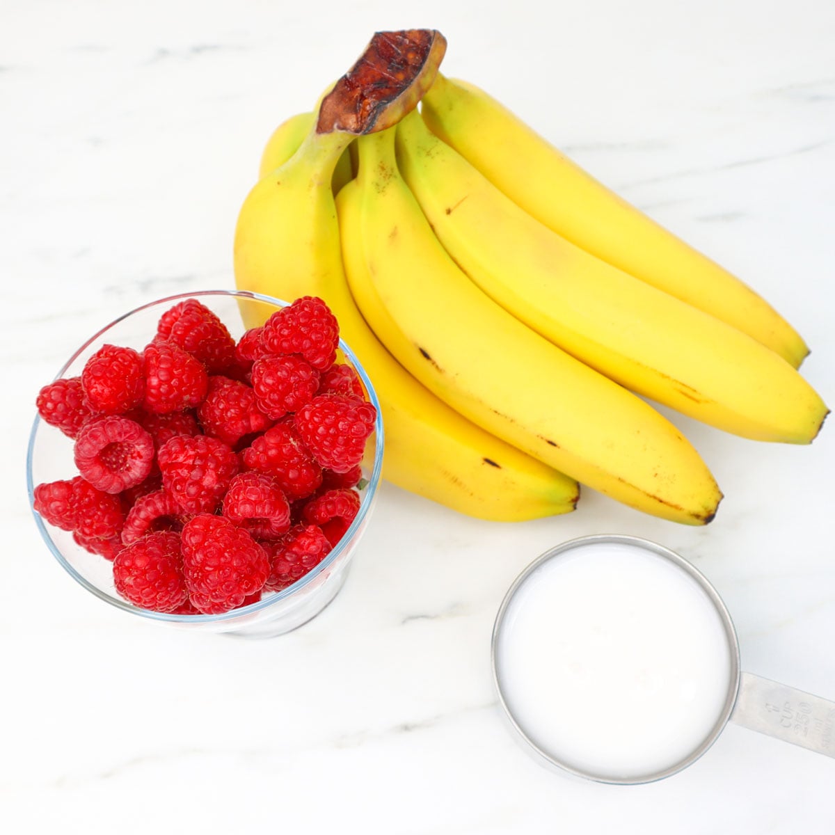 Raspberry Banana Smoothie Freezer Meal Prep Packs A bowl of fresh raspberries next to a bunch of ripe bananas and a measuring cup of yogurt.