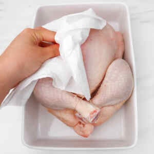 A hand patting a raw chicken with paper towels in a dish.