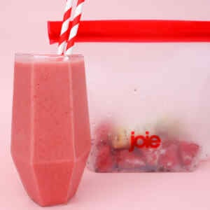 A glass of strawberry-banana smoothie with red and white straws next to a freezer meal prep bag.