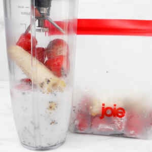 A blender filled with strawberries, banana, chia seeds, and milk next to a freezer bag of frozen fruit.