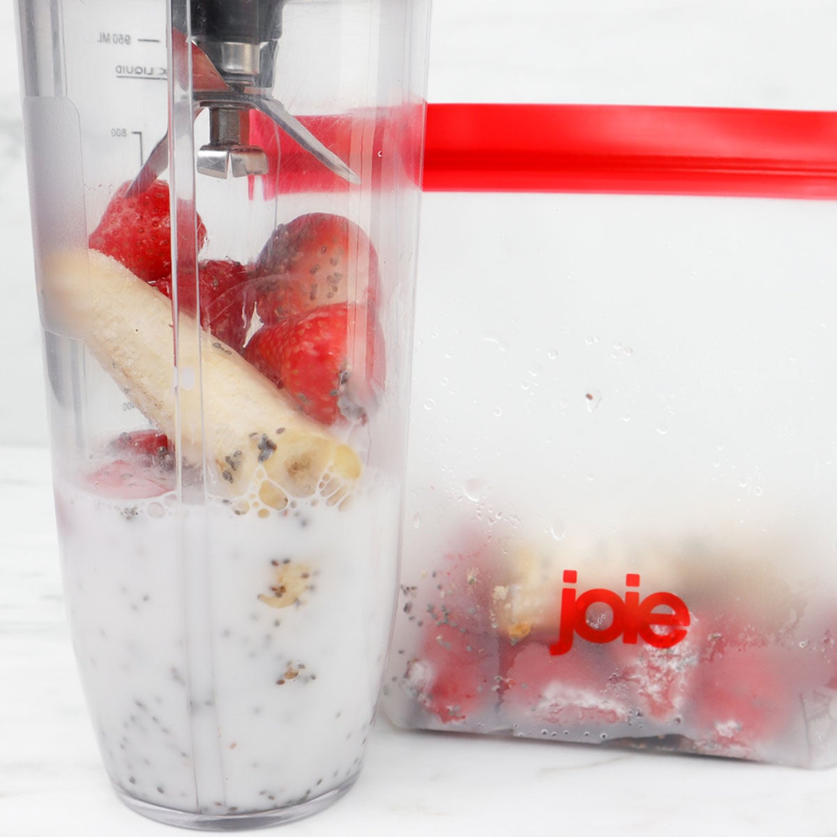 Strawberry and Banana Smoothie Freezer Meal Prep Packs A blender filled with strawberries, banana, chia seeds, and milk next to a freezer bag of frozen fruit.
