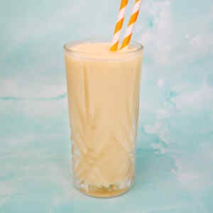 A creamy tropical smoothie in a tall glass with orange and white striped straws.