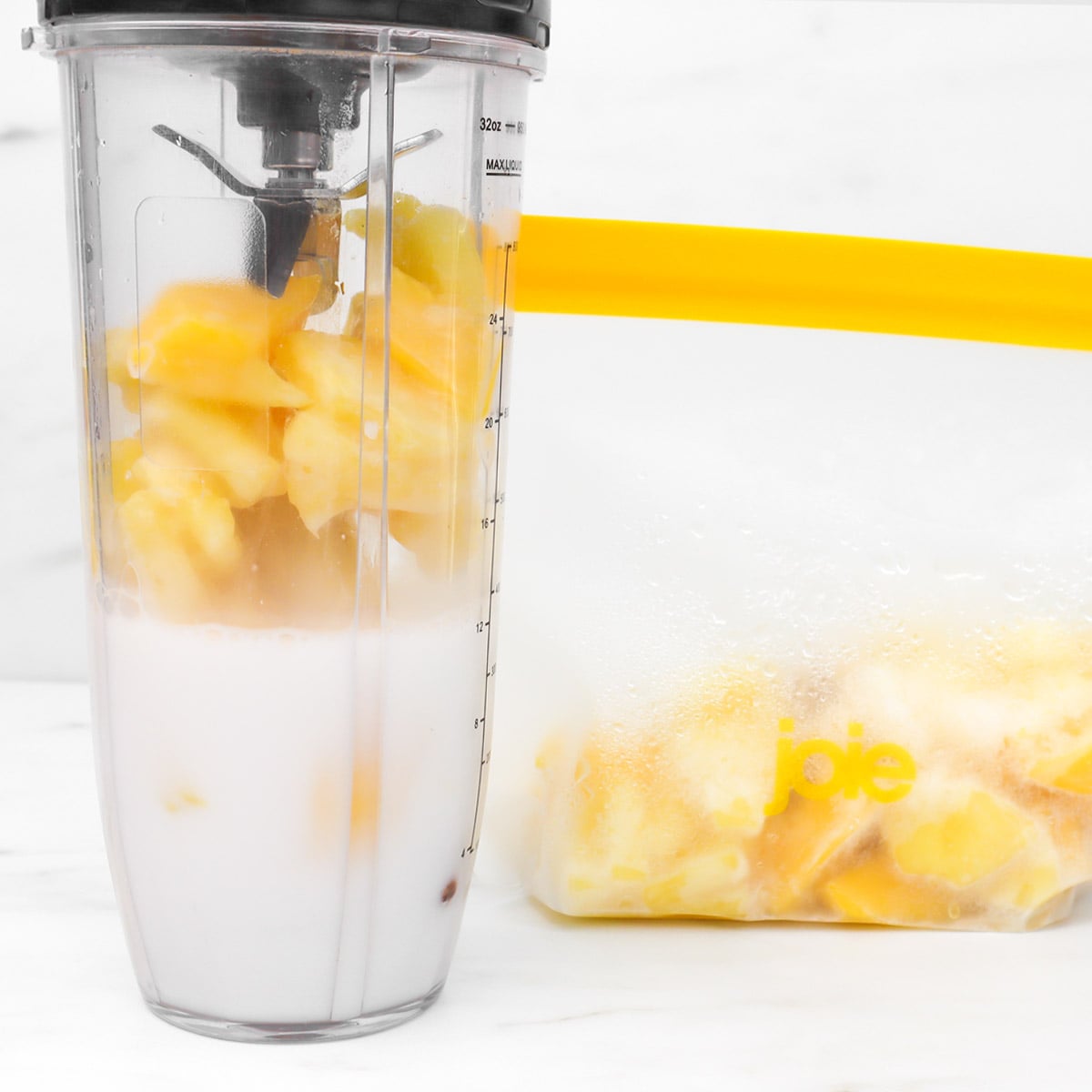 Tropical Smoothie Freezer Meal Prep Packs A blender filled with frozen fruit and milk, next to a bag of frozen fruit.