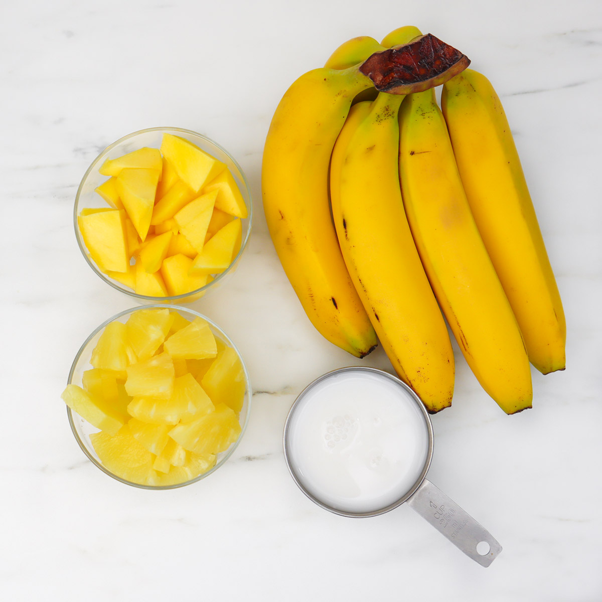 Tropical Smoothie Freezer Meal Prep Packs Ingredients for a tropical smoothie: bananas, pineapple chunks, and coconut milk on a marble surface.