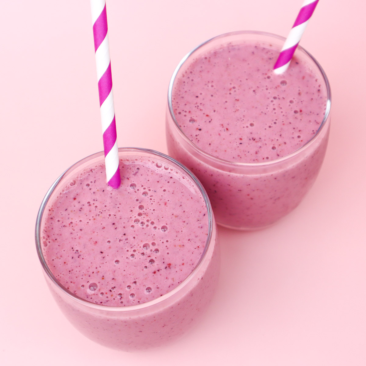 Paquetes de preparación de comidas congeladas con batidos de frutos rojos Dos vasos de batido de frutos rojos con pajitas a rayas rosas y blancas sobre un fondo rosa.