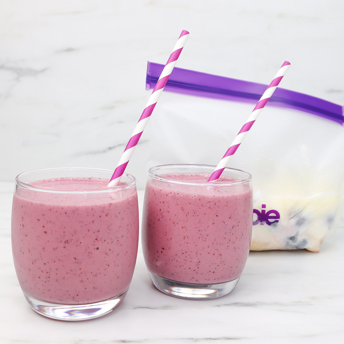 Paquetes de preparación de comidas congeladas con batidos de frutos rojos Dos vasos de batido de frutos rojos con pajitas a rayas y una bolsa de congelación al fondo.
