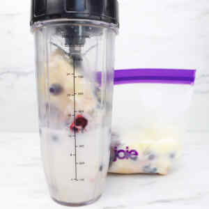 Una batidora llena de ingredientes para batidos junto a una bolsa congelada con frutos rojos variados.