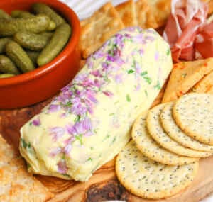 A log of homemade chive flower butter surrounded by crackers, cheese, and pickles.