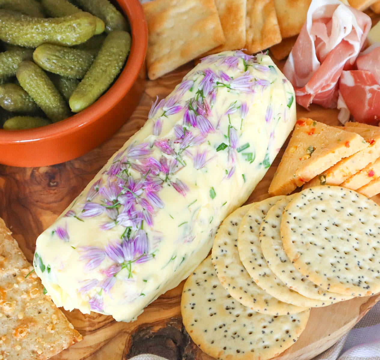 A log of homemade chive flower butter garnished with chive flowers, surrounded by crackers and pickles.